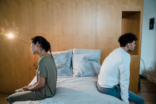 Un uomo e una donna siedono sul bordo del letto dandosi le spalle, visibilmente distanti e privi di intimità fisica ed emotiva.